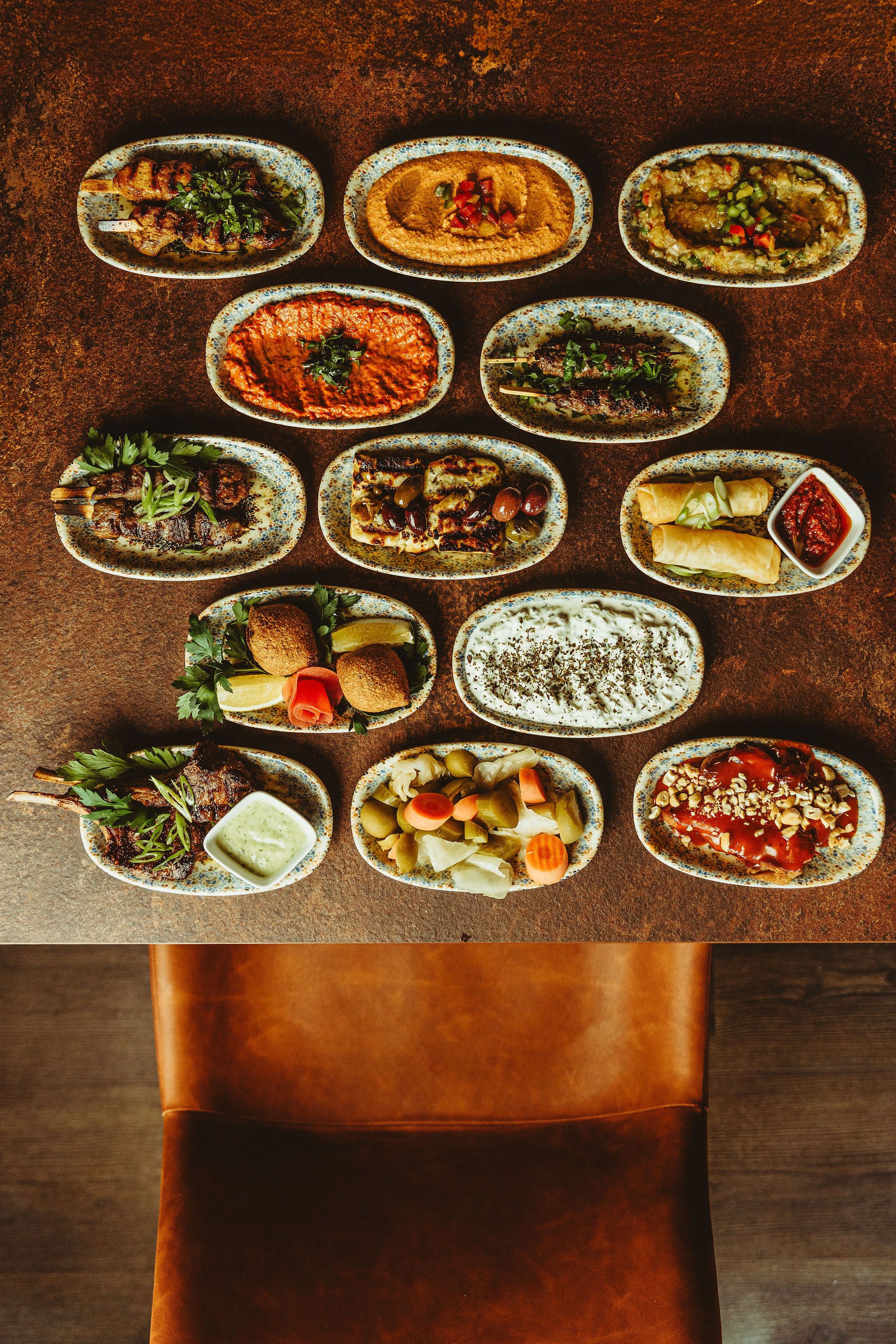 Et bord dækket fra kant til kant med små mezze-retter på mønstrede tallerkener, set ovenfra i varmt aftenlys.
