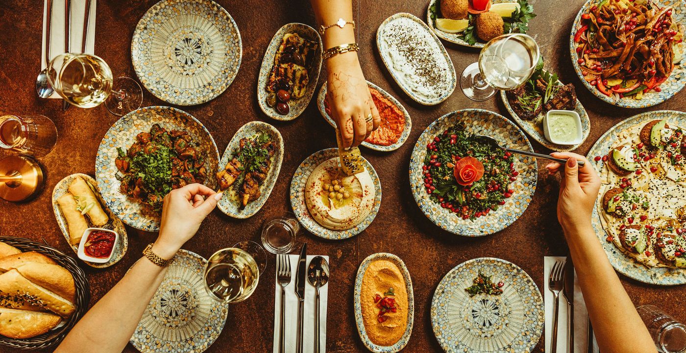 Flere hænder rækker ud over et bord fyldt med mezzeretter, dypper brød i hummus og tager en kødspyd, med en kurv brød i billedets kant.