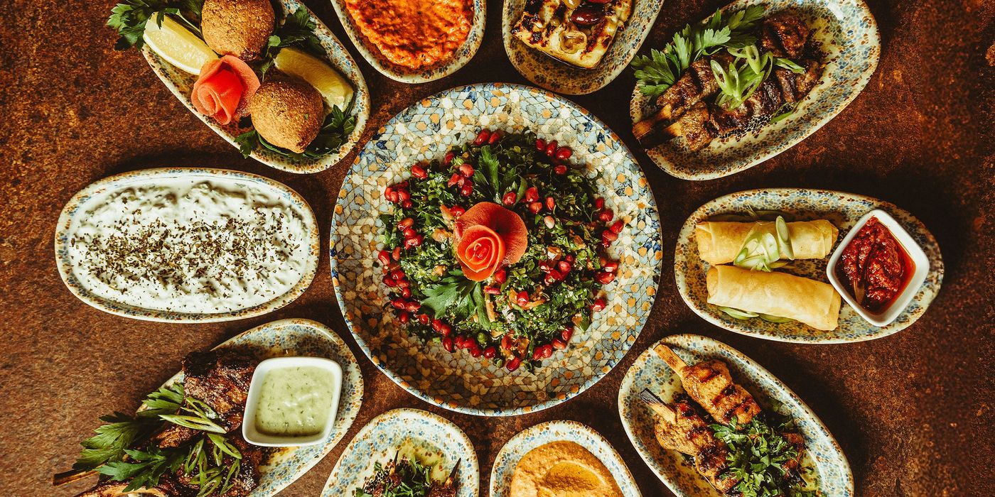 Et bord dækket med mezzeretter arrangeret i en cirkel som en blomst omkring en skål tabbouleh med granatæble og en rose skåret af tomat og gulerod.