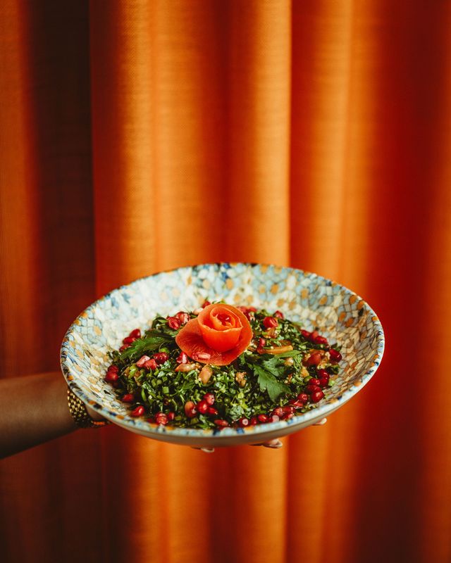 En skål tabbouleh med granatæblekerner holdt frem foran et terracottafarvet gardin.