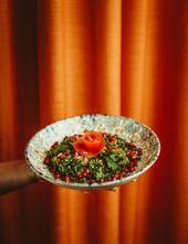 En hånd holder en skål tabbouleh med granatæble og en tomatrose foran et terrakottafarvet gardin.